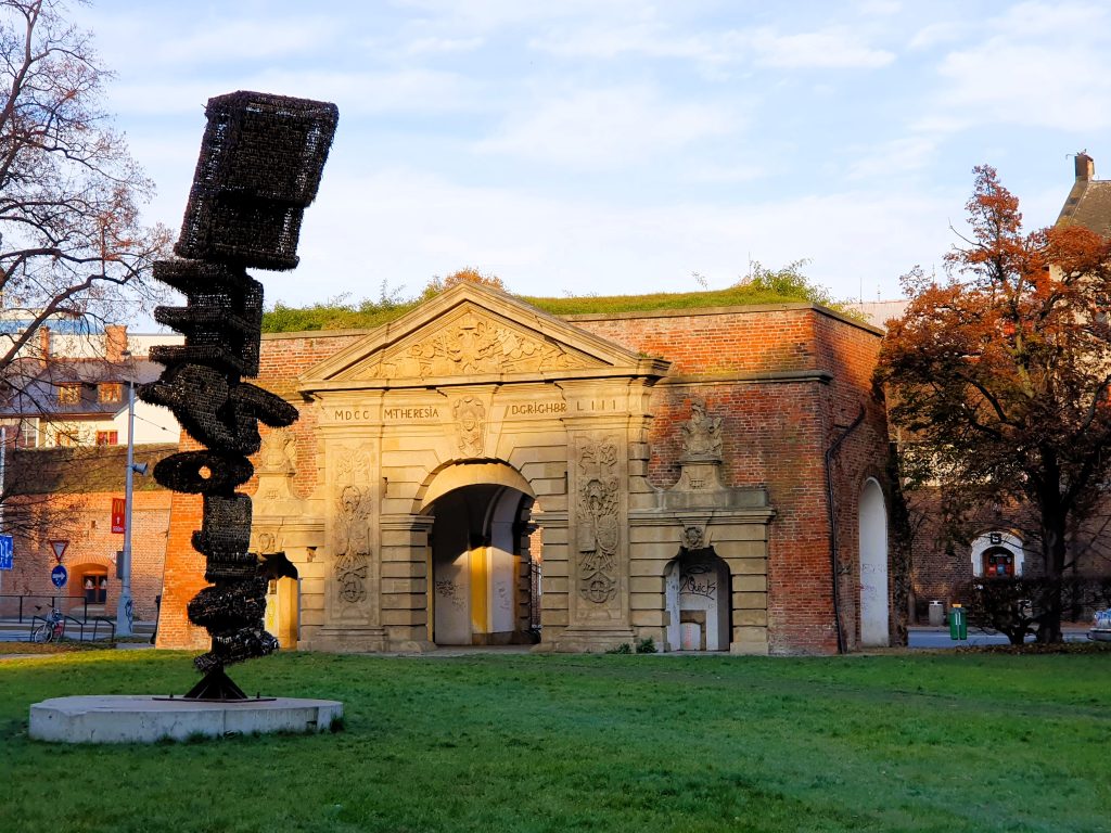 ein historisches Gebäude mit Tor, davor eine moderne hohe Skulptur