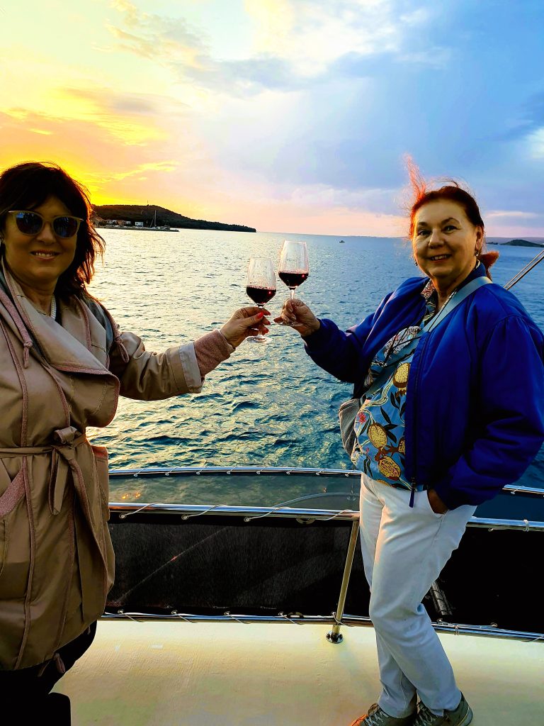 zwei Frauen stehen bei Sonnenuntergang auf Schiffsdeck und trinken ein Glas Wein